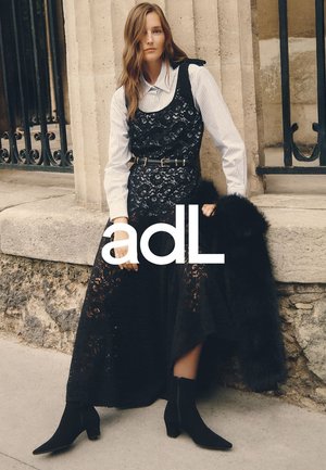 Langes schwarzes Kleid mit Spitzenapplikationen, über einem gestreiften weißen Hemd getragen. Schwarze Knöchelstiefel und ein Pelzakzent vervollständigen den Look.