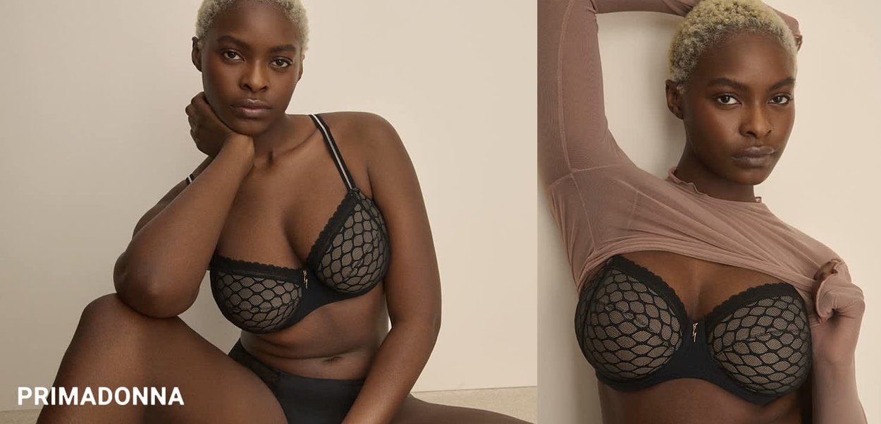 Black lace bra with fishnet pattern and underwire support. Model wears a sheer, light brown top. Smooth skin tone, soft blonde hairstyle.