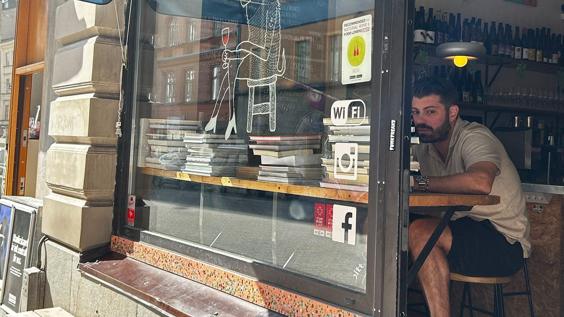 Een café-venster met gestapelde tijdschriften, een houten plank en een tekening van een persoon met een drankje. Nabij zijn sociale media-logo's zichtbaar.