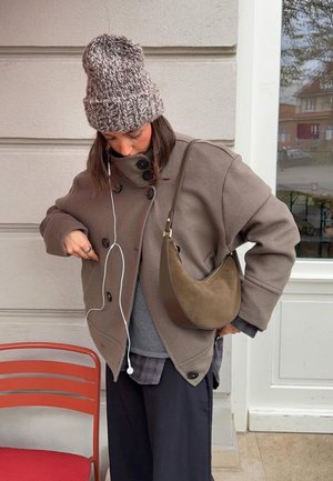 Brauner Wollmantel mit großen Knöpfen, darunter ein grauer Pullover, und eine braune Leder-Umhängetasche. Trägt eine gestrickte Mütze und Kopfhörer.