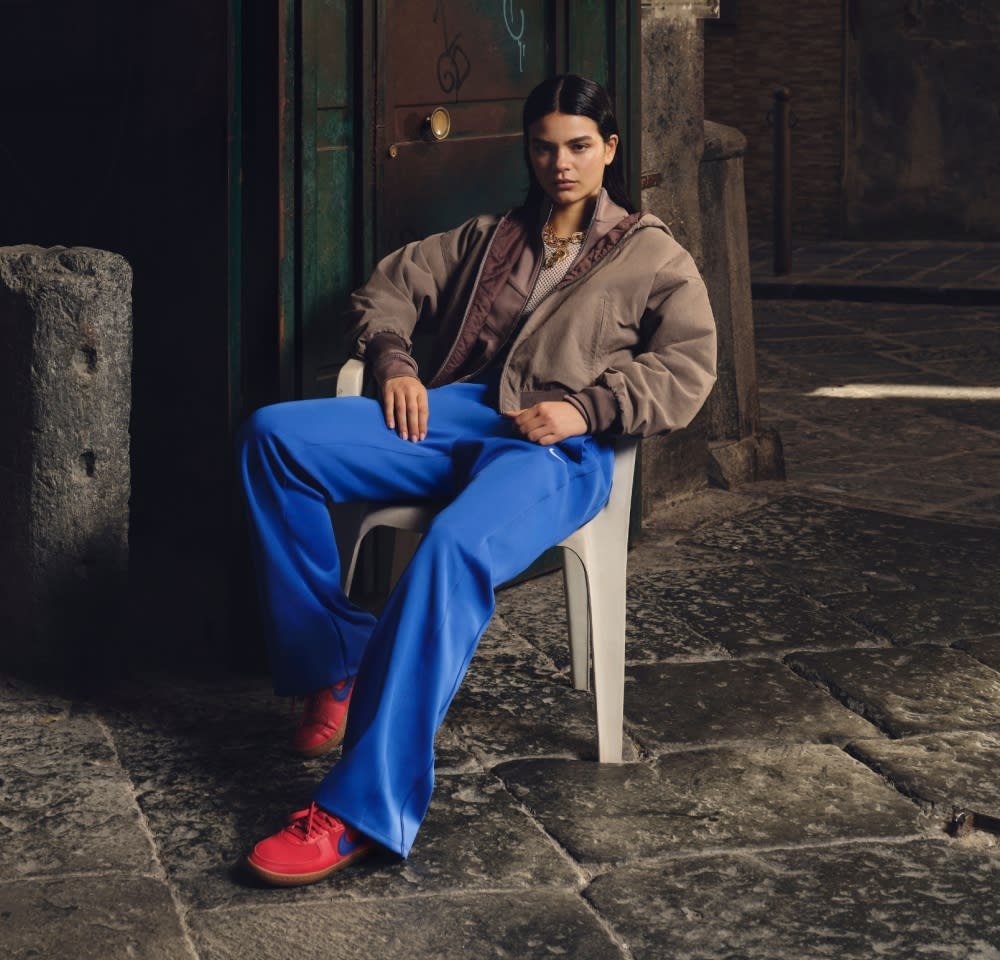 Outfit casual con una giacca bomber marrone, pantaloni larghi blu e sneakers rosse. Il modello è seduto su una sedia di plastica bianca.