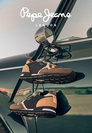 Sneaker in marrone, verde e beige con un mix di suede e tessuto; presenta accenti bianchi e una suola in gomma testurizzata, appesi a uno specchio.