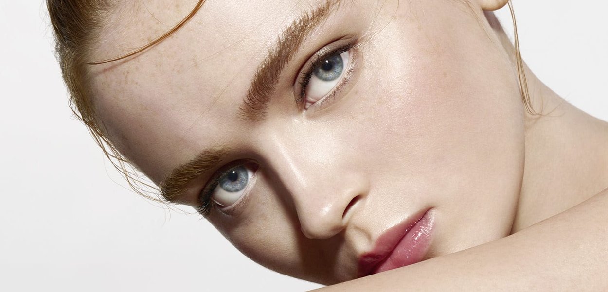 Close-up of a face with smooth, fair skin, light blue eyes, natural brows, and glossy pink lips. Hair is pulled back, revealing skin texture.