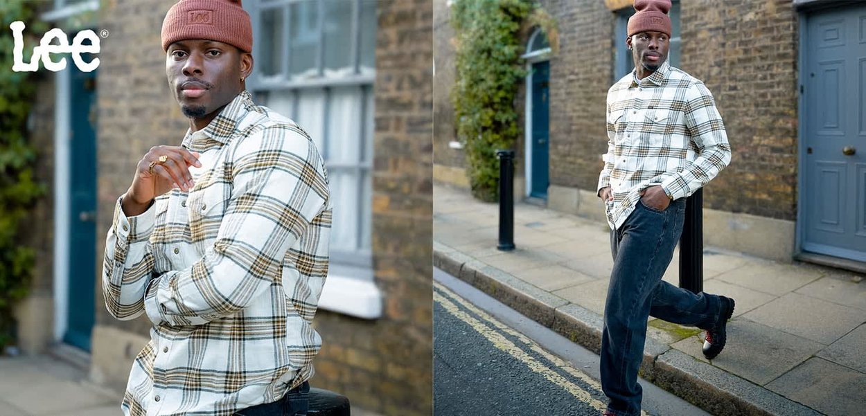 Rutig skjorta i vitt, beige och svart; blå jeans; brun mössa; står på en trottoar nära en röd tegelvägg och en dörr.