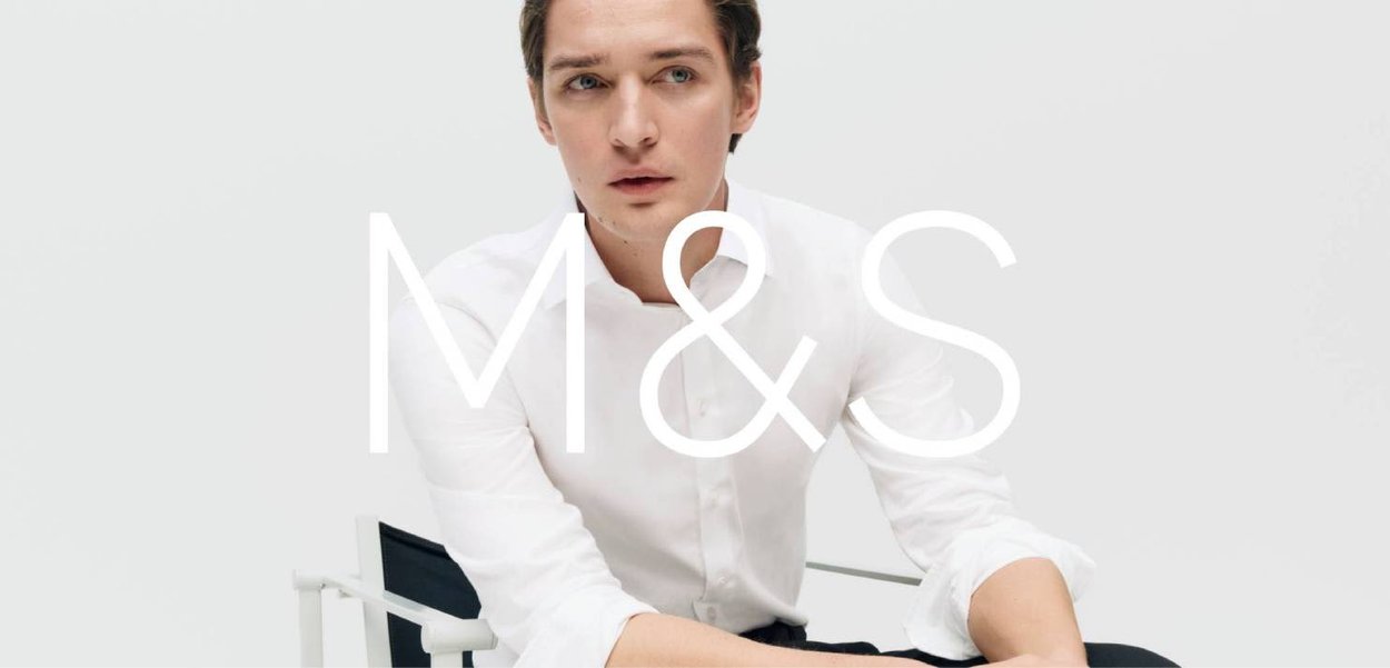 Jeune homme en chemise blanche assis sur une chaise, regardant sur le côté, avec un fond gris clair neutre et une superposition de marque M&S.