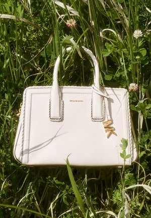 Witte Michael Kors handtas met gouden MK-charme ligt op groen gras met kleine roze bloemen in natuurlijk licht.