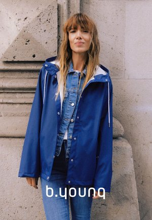 Frau mit langen Haaren lehnt an einer Steinmauer und trägt eine blaue Regenjacke über einer Jeansjacke und Jeans.
