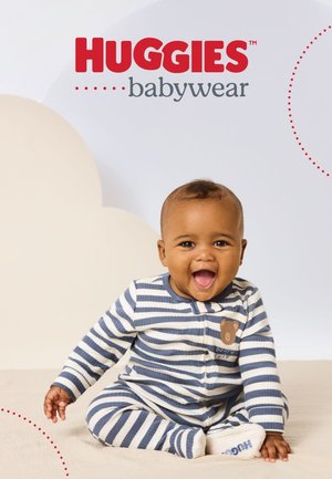 Lächendes Baby sitzt, trägt blaue und weiße gestreifte Huggies-Fußjämmis mit einem Teddybär-Patch auf der Brust, neutraler Hintergrund.