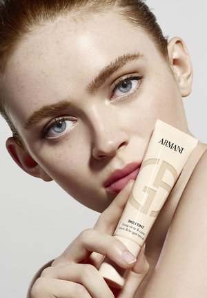Beige tube with clear label reading "ARMANI SKIN TINT" held by a hand. Model with fair skin, light blue eyes, and natural makeup poses.