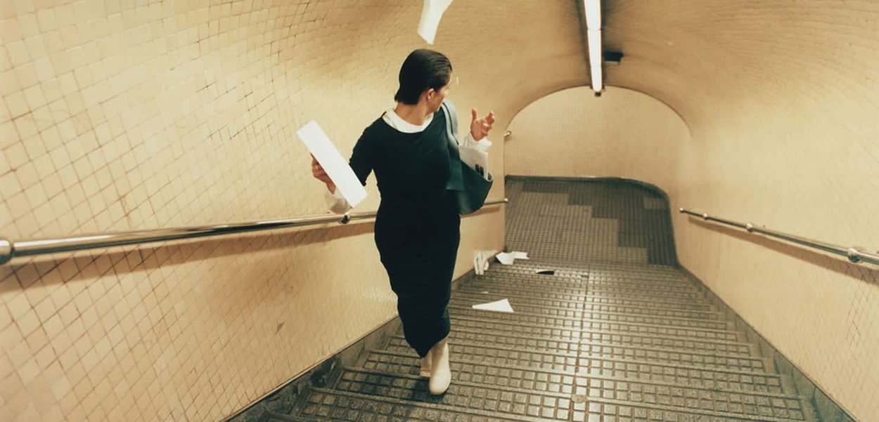 Woman holding papers and walking down a tiled stairwell littered with paper airplanes, wearing black dress and white boots.