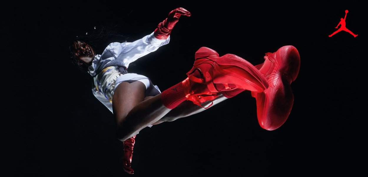 Athlète en tenue de sport blanche et baskets rouges en plein saut, vu de dessous sur un fond noir avec un logo Jumpman rouge dans un coin.