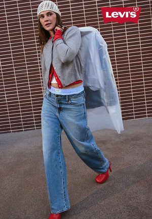 Frau in weiten blauen Jeans, roten Stiefeln, grau-roten Schichten-Oberteilen, weißer Strickmütze, eine Jacke über der Schulter tragend, vor einer gefliesten Wand.