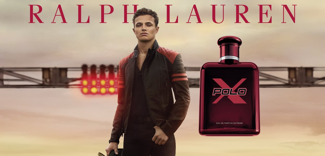 Mann in schwarzer und roter Jacke hält Helm neben einer Rennstrecke, neben ihm steht eine große rote Ralph Lauren Polo X Parfümflasche.