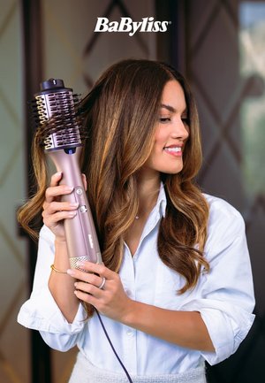 Brosse à cheveux volumisante BaByliss de couleur rose clair, de forme arrondie, avec un design de poils et une prise en main ergonomique. Le modèle la tient tout en coiffant ses cheveux.