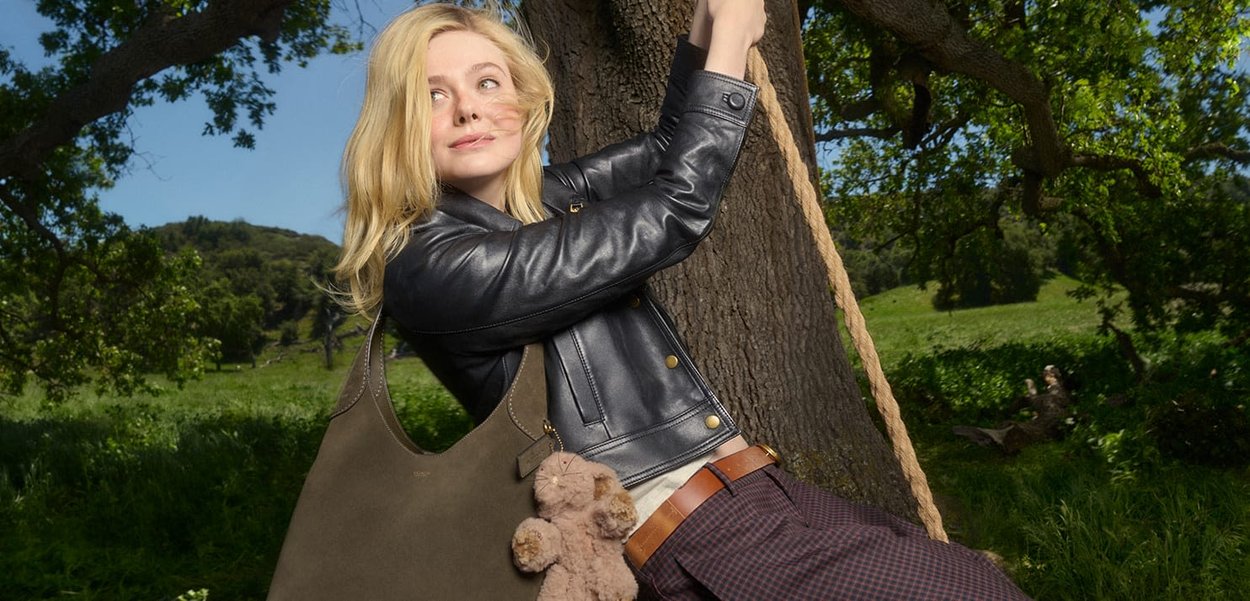 Olive green leather shoulder bag with a soft texture, featuring a plush teddy bear keychain. Model poses against a tree in a natural setting.