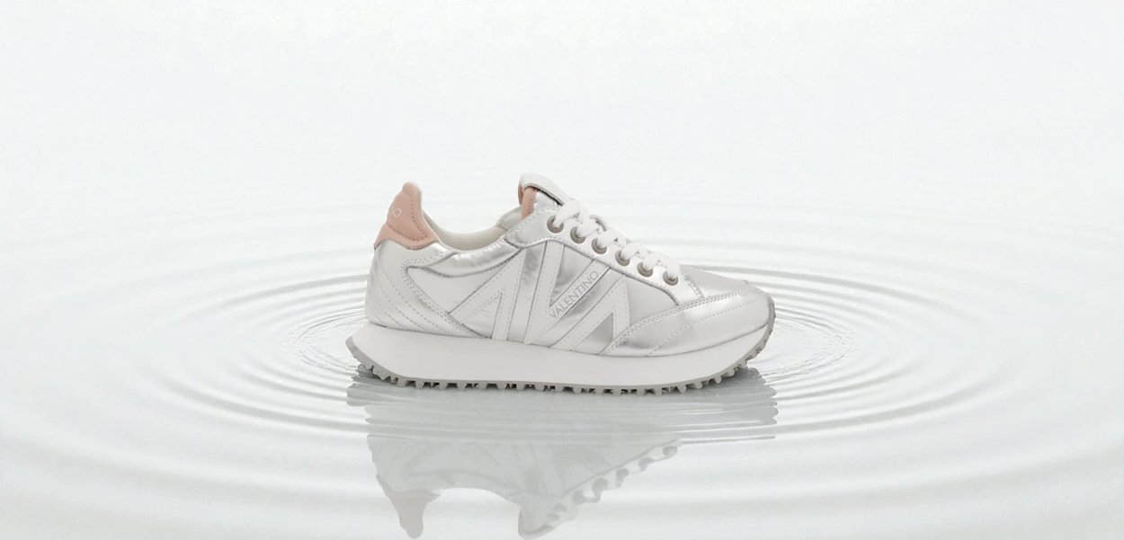 Sneaker sintetica bianca con sottili accenti rosa, pannelli laterali testurizzati, punta rotonda e suola esterna testurizzata, mostrata su una superficie d'acqua increspata.