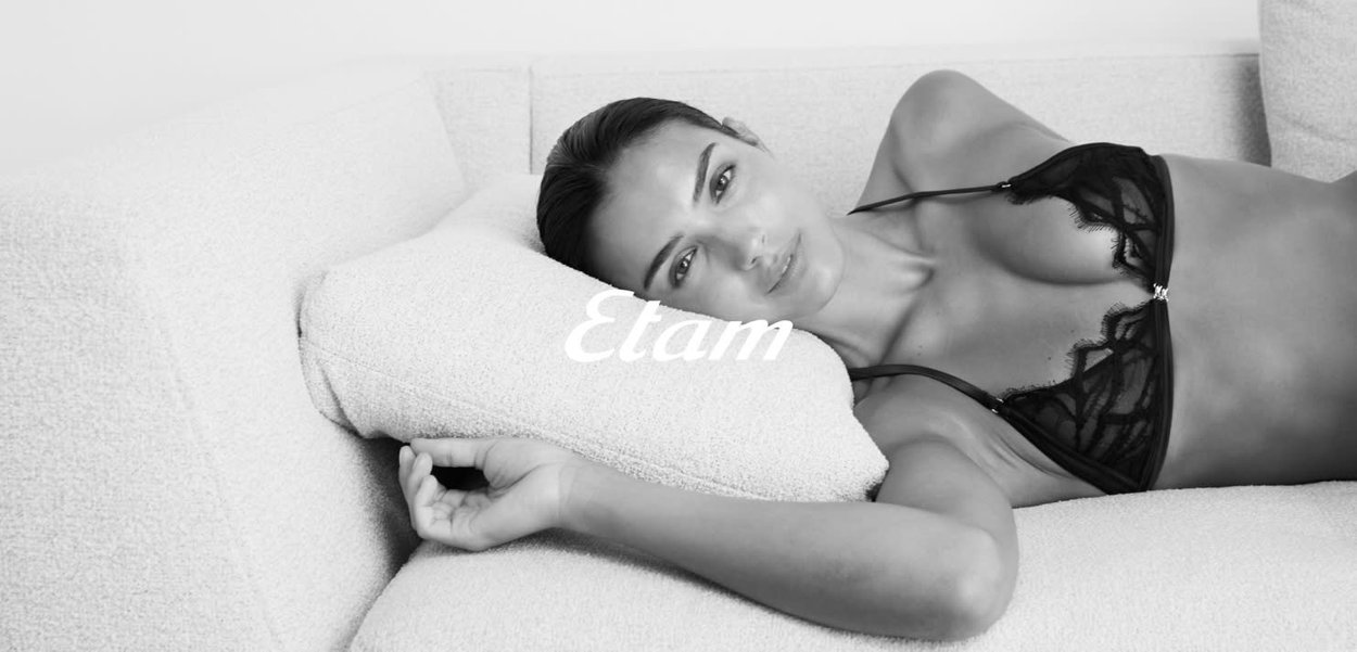 Soutien-gorge en dentelle noire avec des motifs floraux délicats et des bretelles réglables, posé sur un canapé clair et texturé. La marque "Etam" est bien mise en avant.