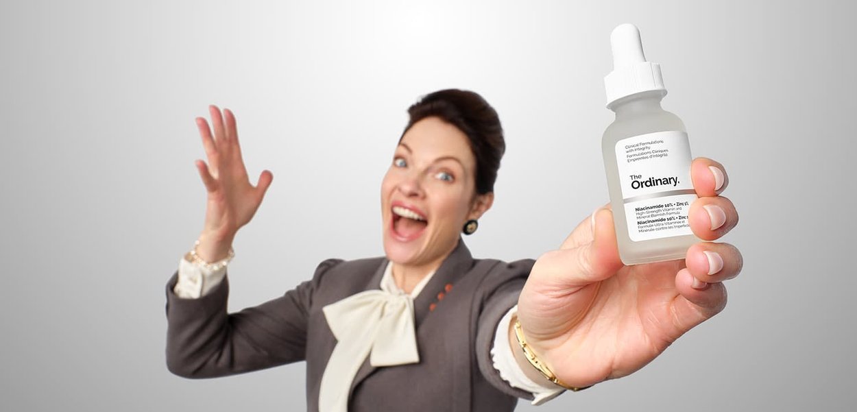 Frau in Businesskleidung, die begeistert eine Flasche des Hautpflegeprodukts The Ordinary mit Pipettenverschluss hält und präsentiert.