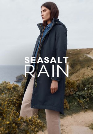 Longue veste de pluie bleu foncé avec capuche, conçue avec une fermeture éclair à l'avant et des accents dorés, portée sur une chemise en denim et un pantalon beige.