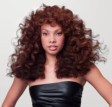 Model with voluminous, curly, auburn hair; wearing a strapless black top. Skin tone is light with freckles. Neutral background.