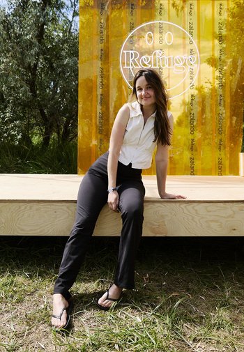 Eine Frau in einer weißen ärmellosen Bluse und schwarzen Hosen sitzt auf einer Holzplattform. Ein durchscheinender orangefarbener Hintergrund zeigt "Refuge" und Markenlogos.