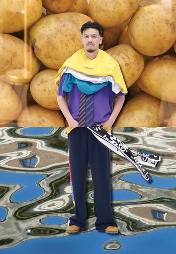 Een persoon staat voor een achtergrond van aardappelen, gekleed in gelaagde kleurrijke shirts, donkere broek en met een patroon sjaal in de hand.