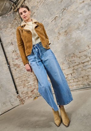 Bruine suède bikerjas, crème kleurige gebreide top, lichtblauwe wijde jeans en tan enkellaarsjes. Gecapitoneerde materialen met een casual ontwerp.