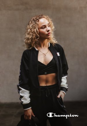 Giacca da track Champion in bianco e nero con zip, design cropped, abbinata a un reggiseno sportivo nero e pantaloni jogger coordinati. Capelli ricci, gioielli minimal.