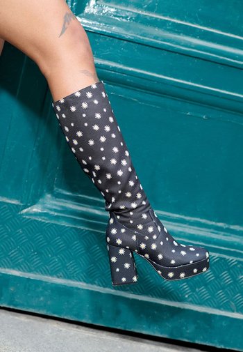 Zwarte kniehoge laarzen met een bloemig madeliefjespatroon, robuuste plateauhak, soepele textuur en ronde neus, tegen een teal achtergrond.