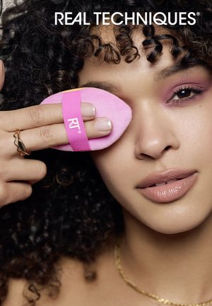 Pink makeupsvamp med rem, holdt i en hånd. Krøllet hår og et lyst, neutralt makeup-look i baggrunden. Lyserøde detaljer synlige.