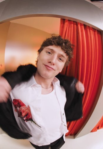 Jeune homme aux cheveux bouclés en train d'enfiler une veste noire, portant une chemise blanche imprimée de roses rouges, debout devant des rideaux orange.