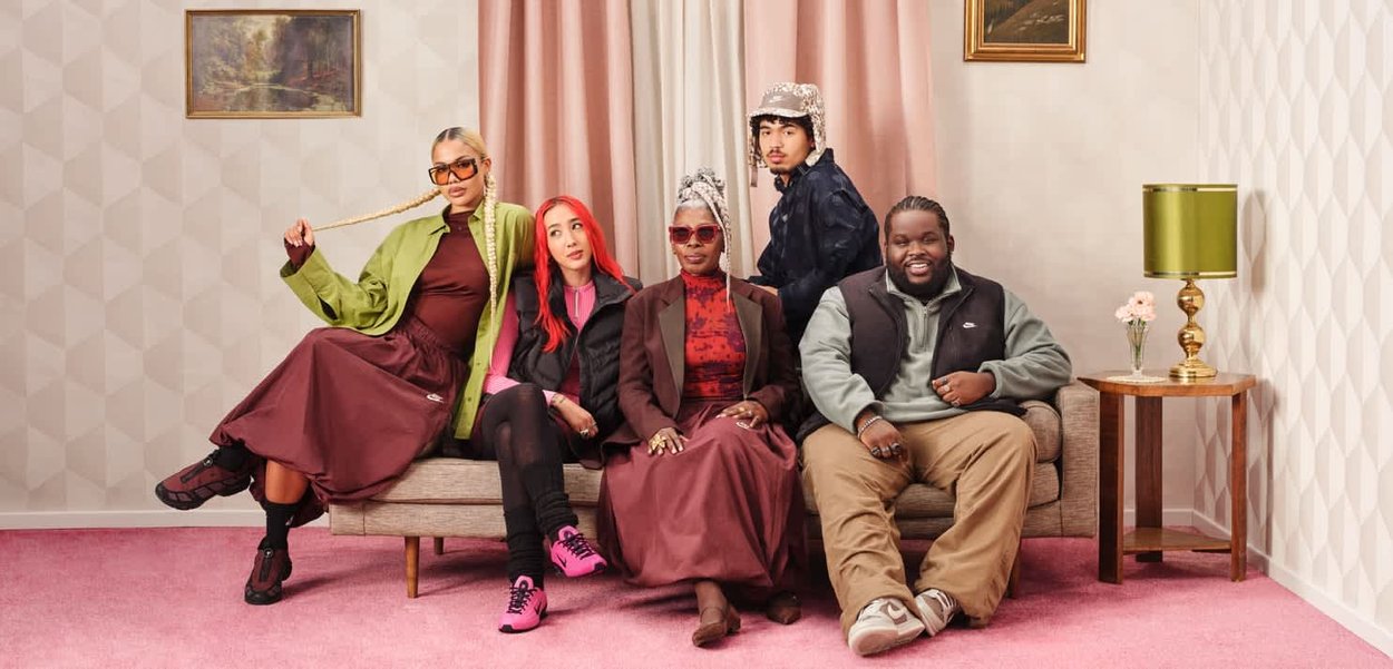 Gruppo di cinque persone sedute su un divano beige. Tappeto rosa con trama, carta da parati a motivi e una lampada verde con fiori nelle vicinanze.