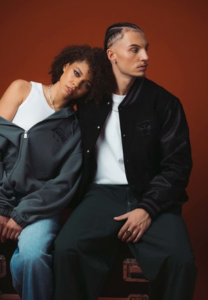 Zwei Personen tragen legere Streetwear. Die Frau hat einen grauen Zip-Hoodie und hellblaue Jeans an, während der Mann eine schwarze Collegejacke und ein weißes Shirt trägt.