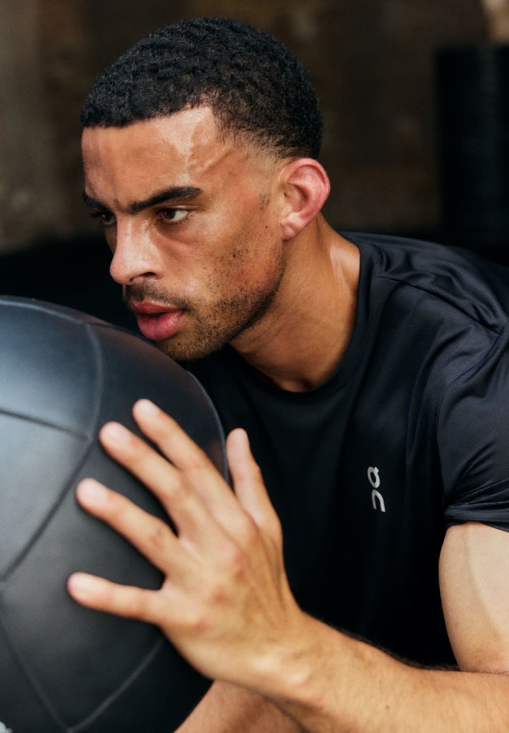 Schwarzes Sportshirt aus feuchtigkeitsableitendem Material. Ein Mann hält einen schwarzen Medizinball, zeigt definierte Armmuskeln und einen konzentrierten Gesichtsausdruck.