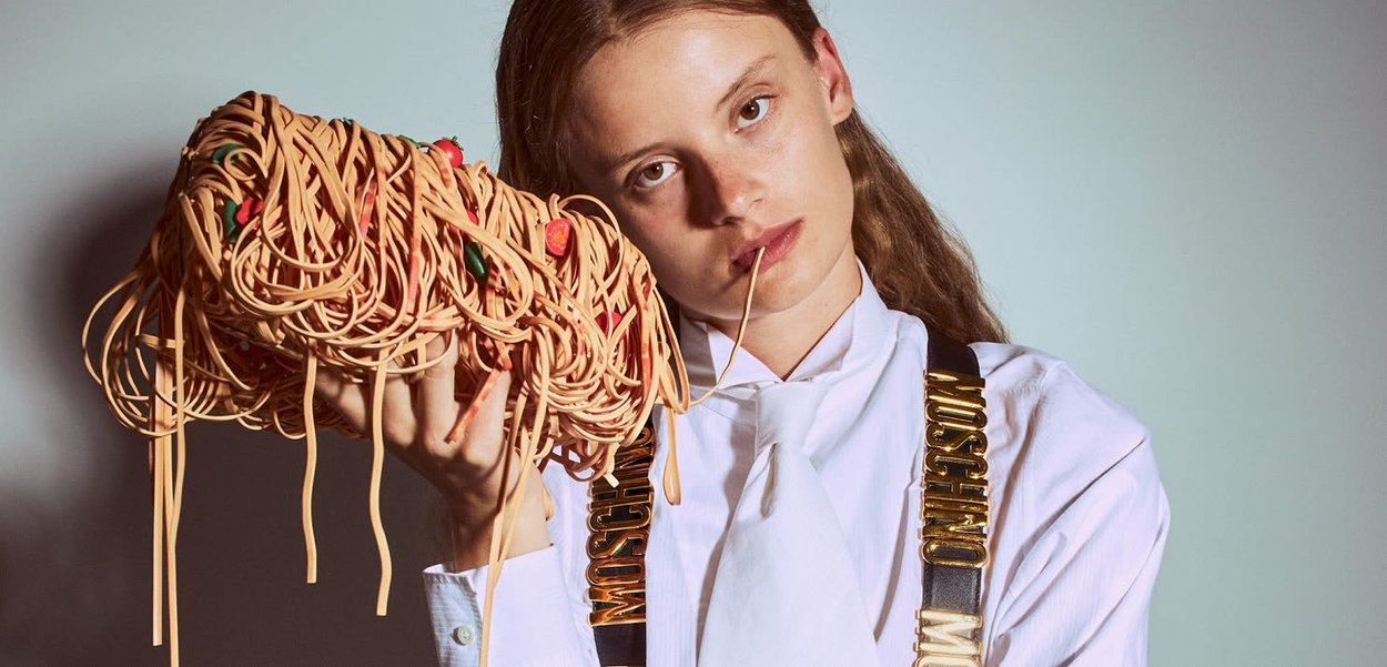 Una modella tiene in mano una borsa a forma di spaghetti, realizzata con materiali color panna, caratterizzata da decorazioni colorate e un design disordinato simile a una pasta.