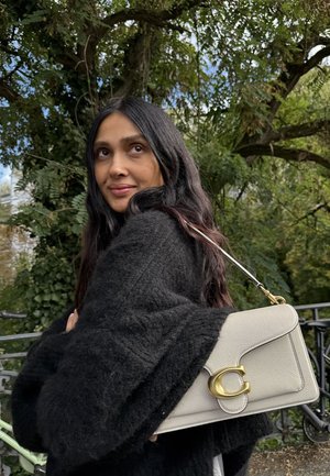 Helle graue Lederhandtasche mit einem goldenen runden Logo, strukturiertem Design und einem abnehmbaren Schultergurt, gehalten gegen einen schwarzen Pullover.