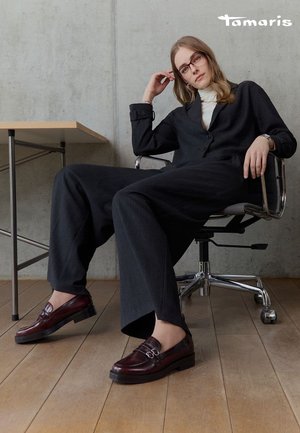 Femme portant un costume noir et un col roulé blanc, assise sur une chaise de bureau, mettant en valeur des mocassins bordeaux foncés sur un sol en bois.