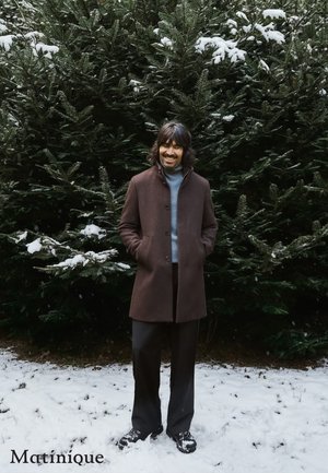 Brown wool coat with a high collar, two side pockets, and button closure, worn over a light blue turtleneck, paired with dark trousers.