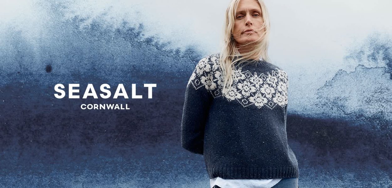 Lyshåret kvinde iført en marineblå mønstret sweater står foran en tekstureret blå væg med teksten "SEASALT CORNWALL".