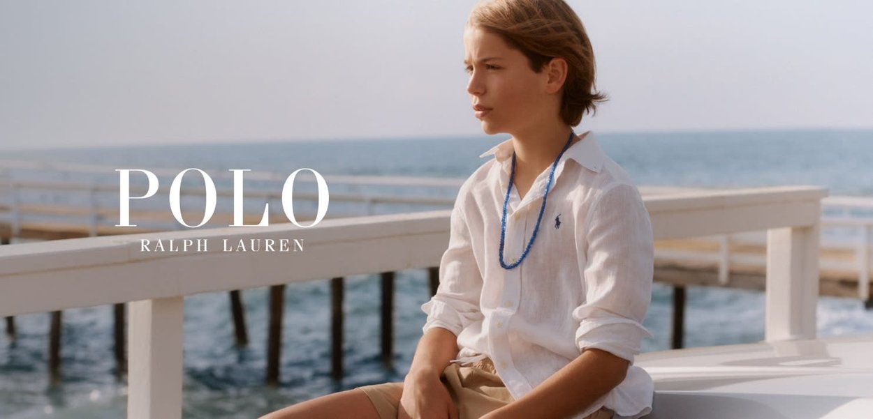 Persona joven con camisa blanca y pantalones cortos beige sentada en una barandilla blanca junto al mar, con el logo de Polo Ralph Lauren visible.