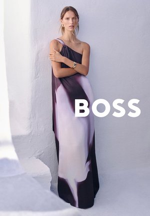 Mujer con un vestido largo de un solo hombro en degradado morado y blanco, de pie contra una pared blanca con textura, llevando una pulsera y pendientes de oro.