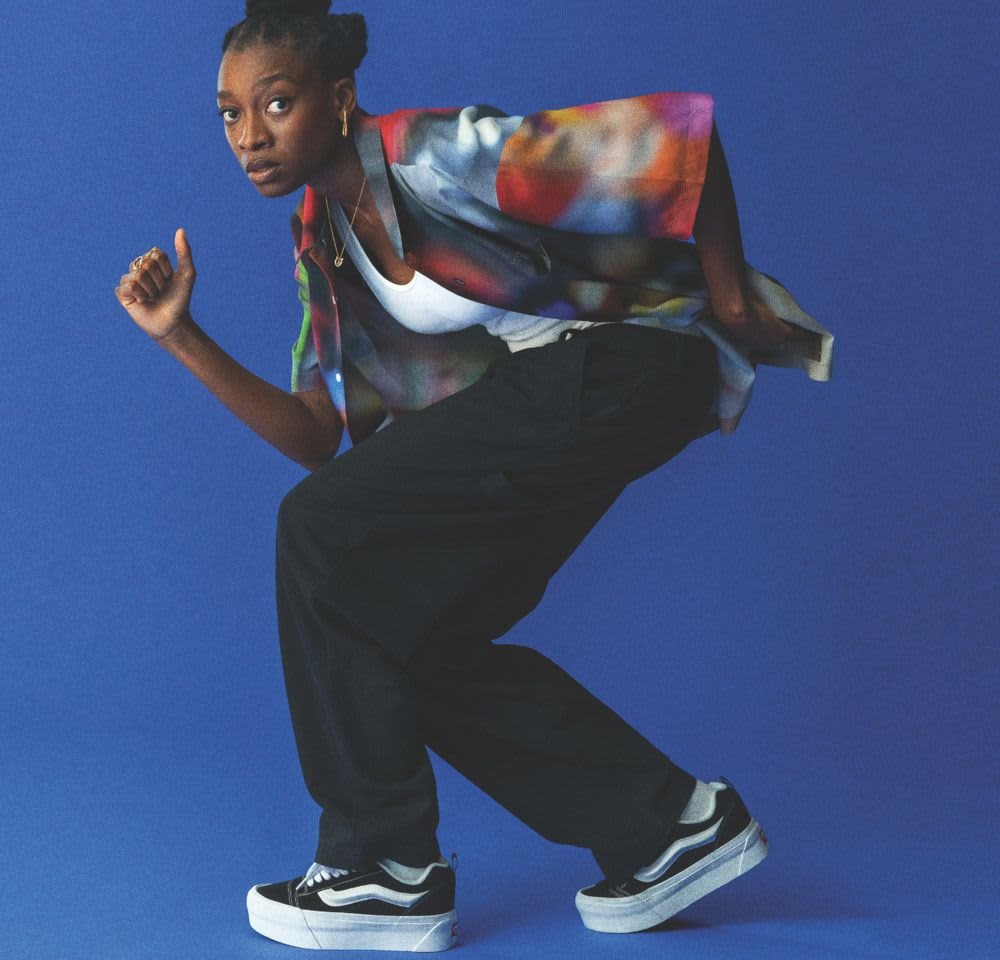 Buntes Kurzarmhemd mit abstrakten Mustern, kombiniert mit schwarzen lockeren Hosen und schwarzen Sneakers mit weißen Sohlen. Der Hintergrund ist blau.