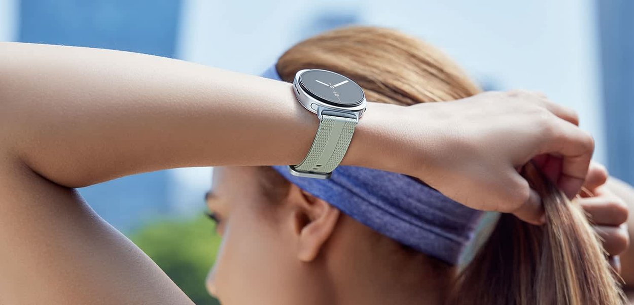 Smartwatch mit schwarzem Zifferblatt und silbernem Rahmen, befestigt an einem hellgrünen strukturierten Armband, das an einem Handgelenk getragen wird, mit einem verschwommenen Outdoor-Hintergrund.
