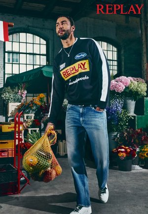 Svart hettegenser med hvite detaljer, stor farget logo, blå jeans og hvite joggesko. Holder en gul nettingpose med frukt. Blomstrende bakgrunn.