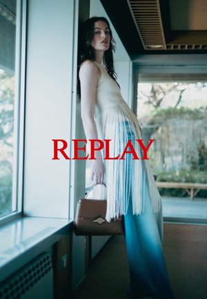 Cremfarbendes Fransenkleid mit fließendem Design, kombiniert mit weit geschnittenen blauen Jeans. Braune Handtasche mit dekorativem Verschluss. Auffälliger roter "REPLAY"-Schriftzug.