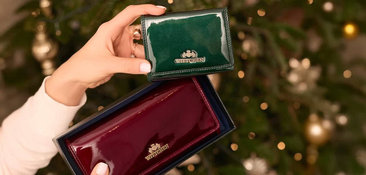 Deux portefeuilles WITTCHEN, l'un vert et l'autre bordeaux, tous deux en cuir brillant, tenus dans une main au-dessus d'une boîte cadeau. Accents de logo doré visibles.