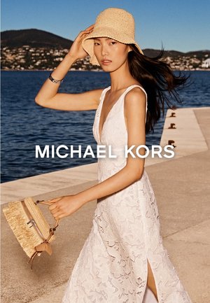 Mujer junto al agua con un vestido blanco de encaje y un sombrero tipo bucket de crochet beige, sosteniendo un bolso de paja en un día soleado en un muelle.
