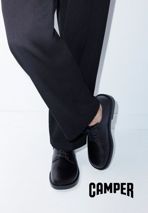 Persona con zapatos de vestir negros y pantalones negros cruzando una pierna sobre la otra, logo de la marca Camper visible.