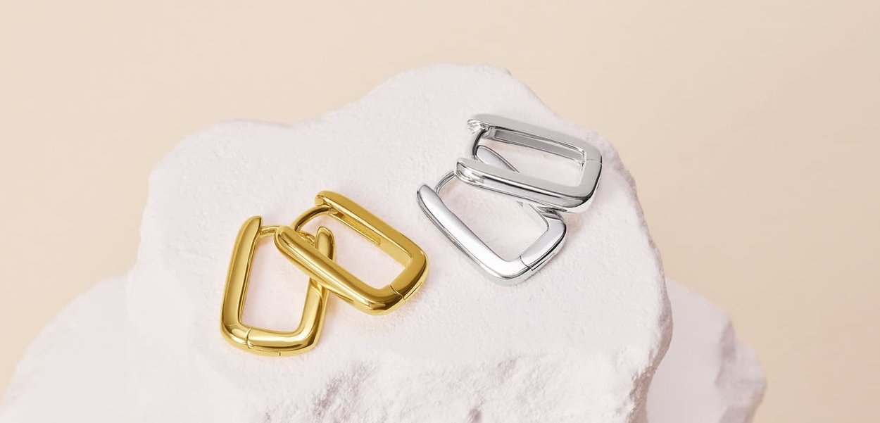 Fibbia in metallo oro e argento, forma rettangolare con bordi arrotondati, superficie lucida, posizionata su uno sfondo di roccia bianca testurizzata.