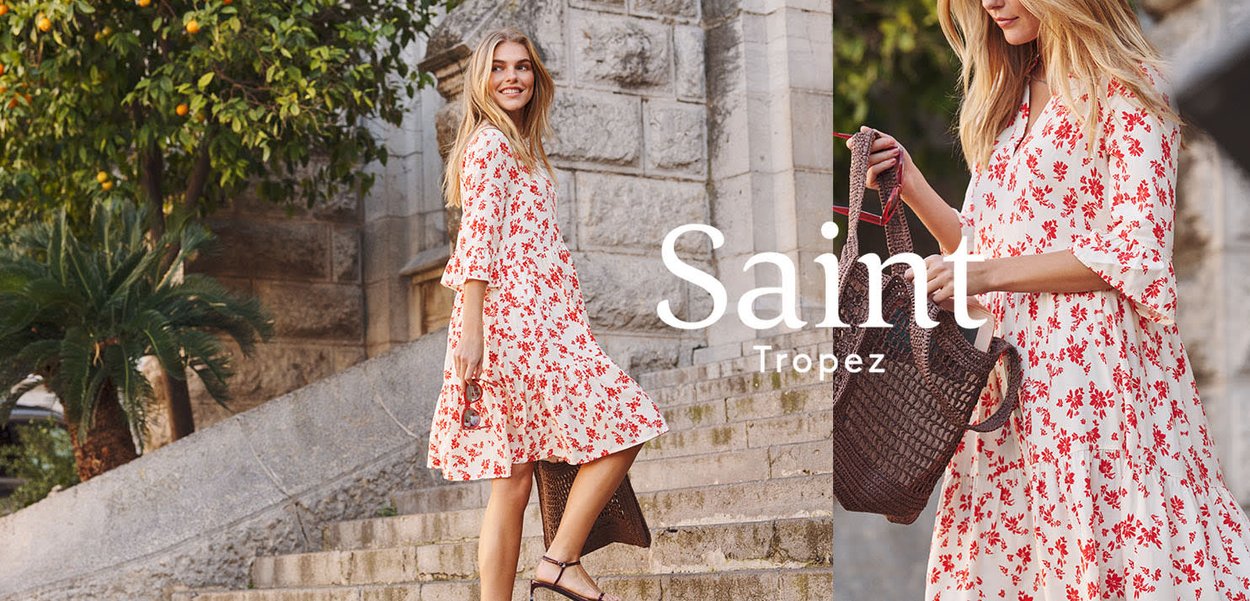 Frau in weißem Kleid mit rotem Blumenmuster, die auf Steinstufen steht und im Freien eine große braune geflochtene Tasche neben grüner Vegetation trägt.